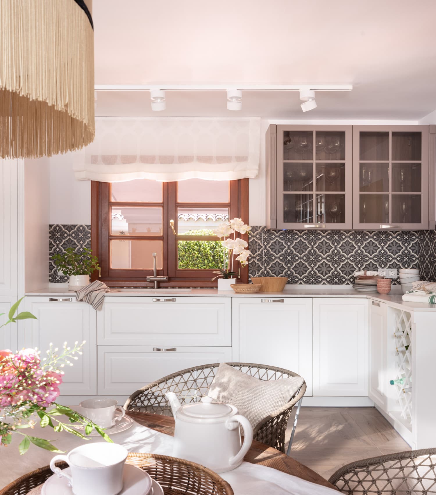 Primer plano de la zona de trabajo de la cocina con armarios bajos en blanco lacado y alacenas superiores en gris topo, separadas por un espectacular zócalo de azulejo hidráulico de motivos florales en negro y blanco. Ventana de madera con estor de gasa y orquídea blanca sobre la encimera. La lámpara de flecos crea un efecto visual cálido sobre la mesa auxiliar.