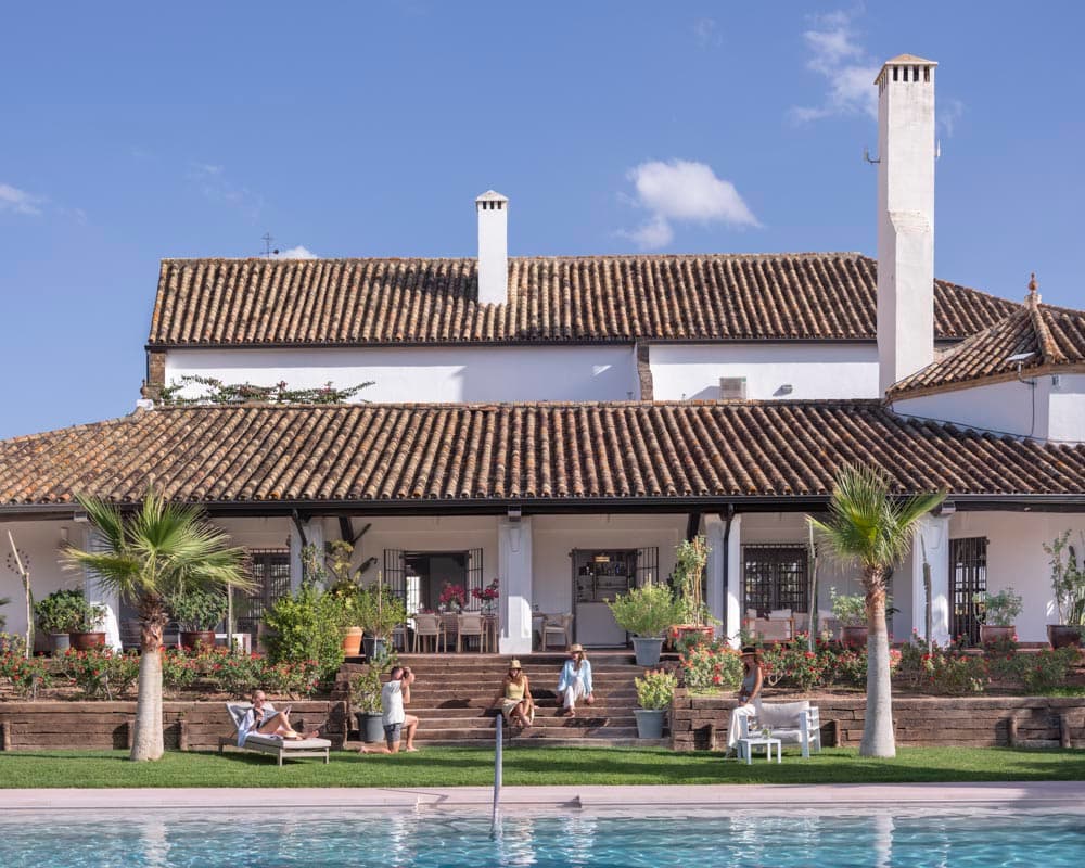 Vista exterior de la casa desde la piscina. Fachada de dos plantas encalada en blanco con tejado de teja árabe y chimeneas, rodeada de palmeras, jardín escalonado y pérgola cubierta con vigas negras.