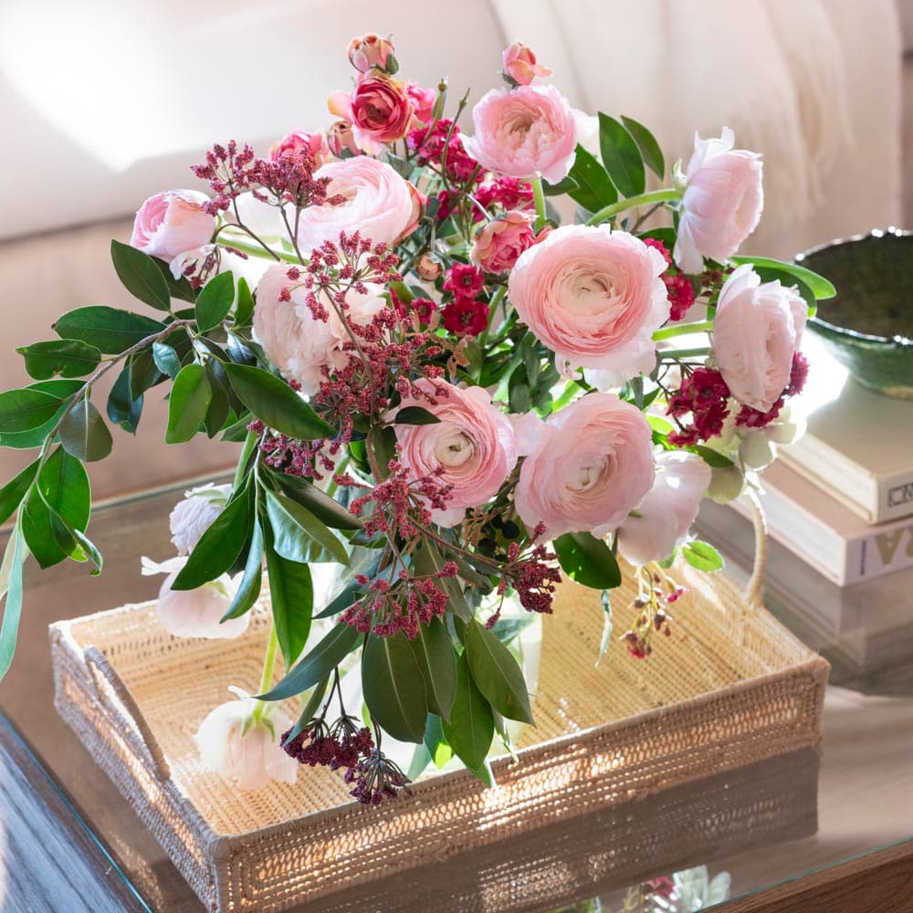 Primer plano de un generoso ramo de ranúnculos rosados, alstroemerias y flores silvestres en vaso de cristal sobre una bandeja rectangular de ratán, colocada sobre una mesa de cristal en el salón. La composición floral aporta delicadeza romántica al ambiente.
