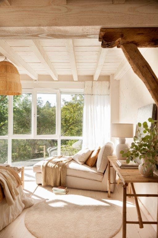 Acogedor rincón de lectura con sofá blanco en lino, viga de madera rústica en diagonal y grandes ventanales con vistas al jardín. Una mesa auxiliar de madera clara y plantas complementan este espacio lleno de luz natural.