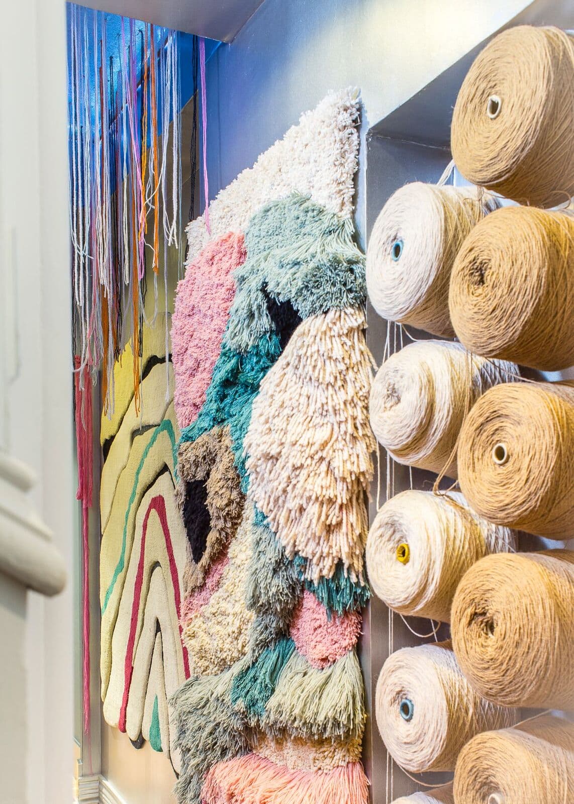 Primer plano de un tapiz de pelo largo con tonos rosas, verdes y crema junto a una estantería con grandes conos de hilo natural, mostrando el proceso creativo y los materiales del estudio de Dafne Vijande.