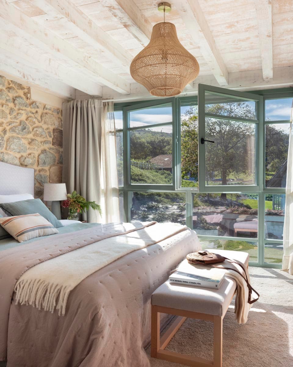 Dormitorio sereno con pared de piedra vista, cama tapizada con ropa de cama en verde agua y beige, gran galería acristalada de marco verde que inunda de luz natural el espacio y una lámpara de ratán tipo farolillo.