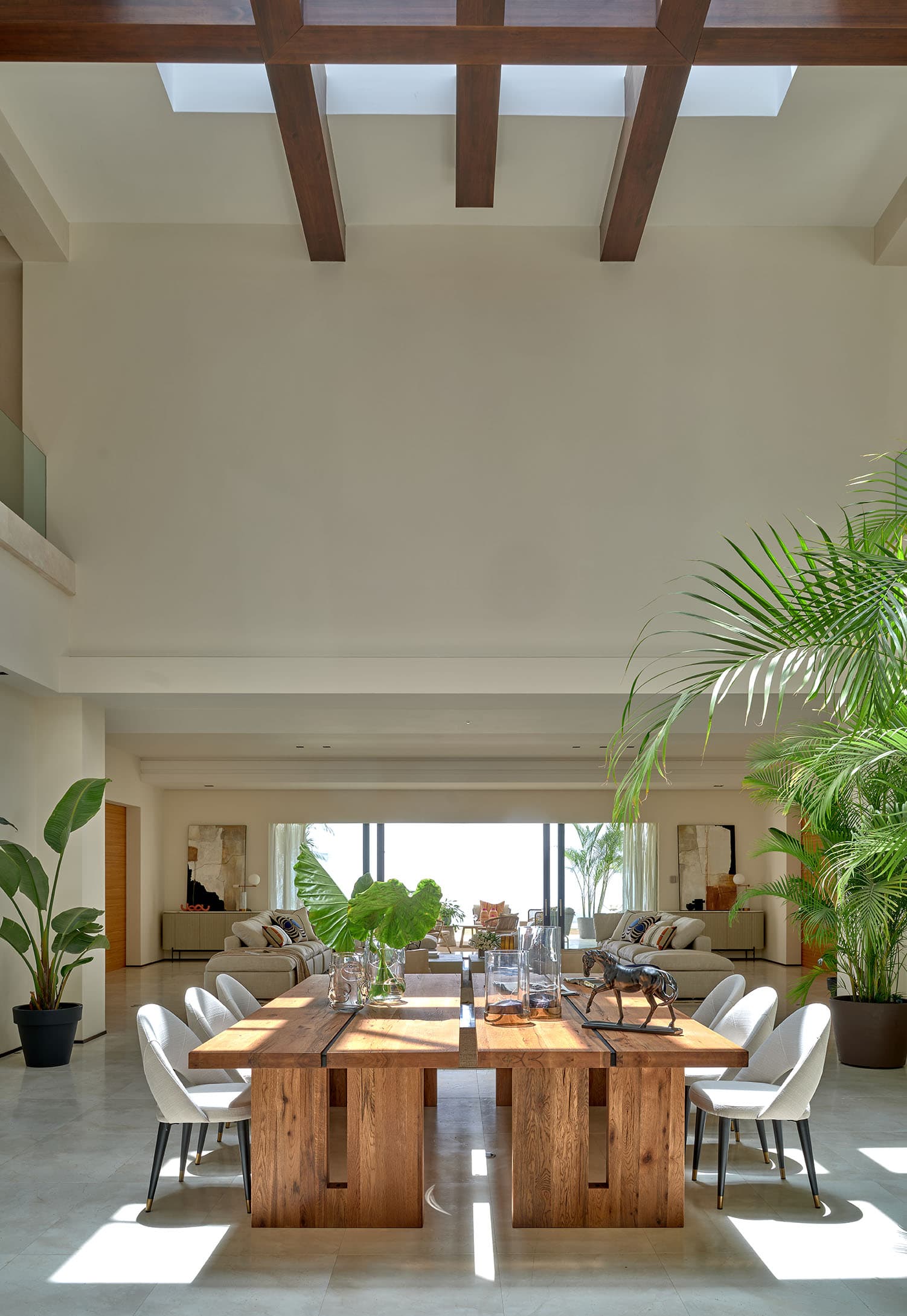 Comedor de doble altura con techo de vigas de madera oscura y lucernario, gran mesa rústica de tablones con sillas de tapizado gris perla y palmeras monumentales en el interior.
