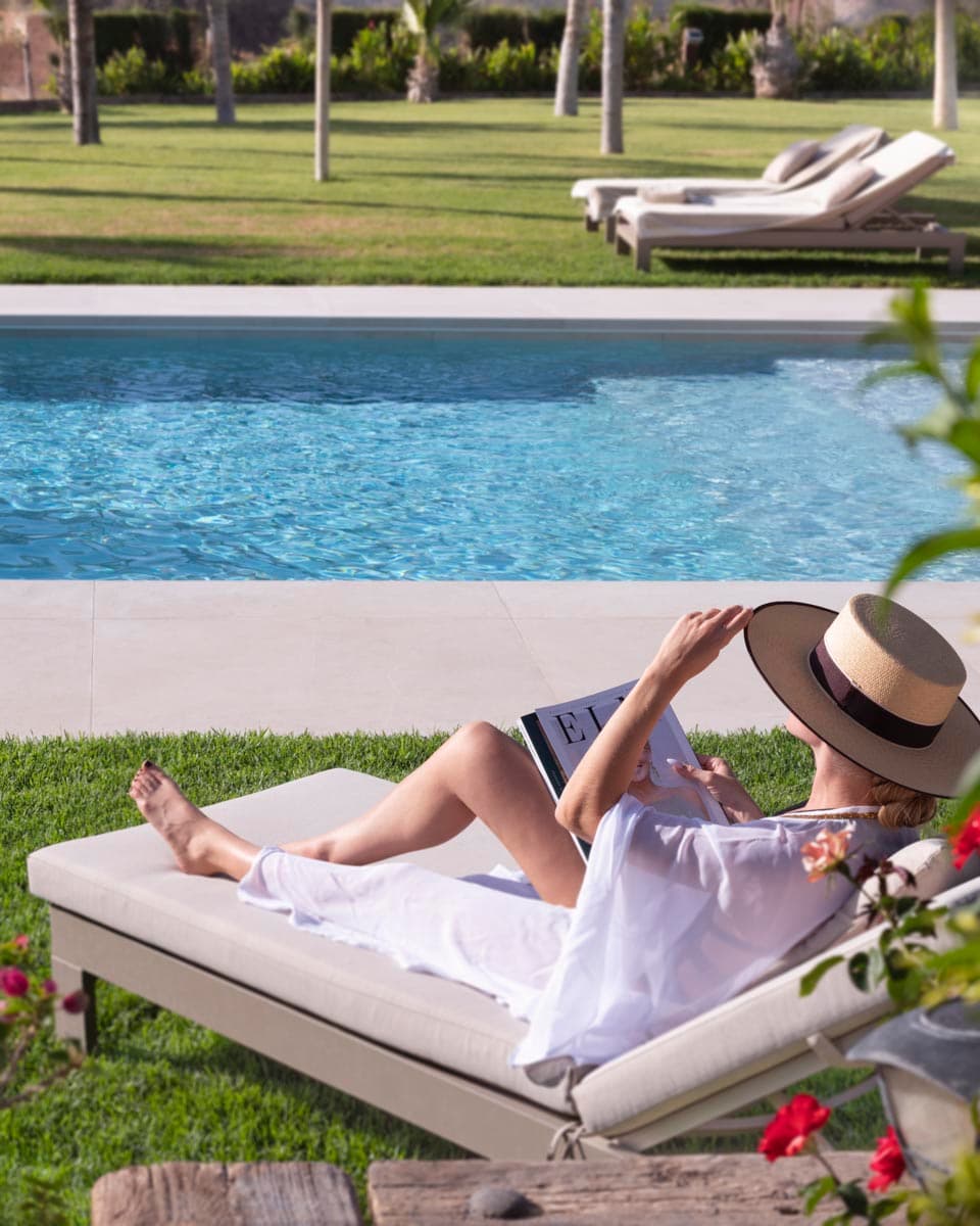 Mujer leyendo una revista recostada en una tumbona beige junto a la piscina de borde infinito. Césped cuidado, flores rojas en primer plano y más tumbonas al fondo con vistas al jardín.