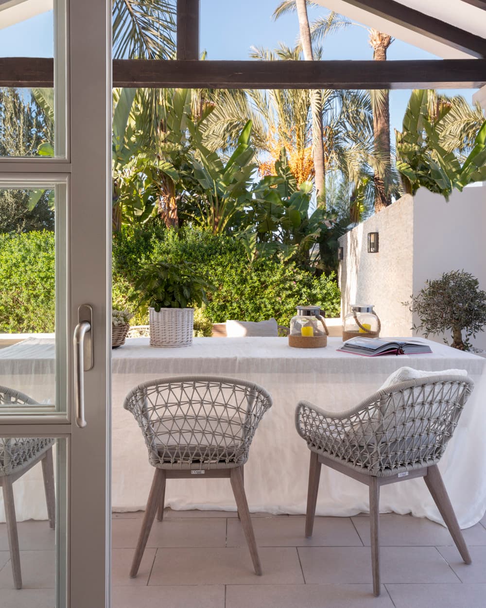 Vista desde las puertas acristaladas de dos sillas de cuerda gris ante la mesa con lino blanco y jarras de cristal, con el exuberante jardín tropical de bananeros y palmeras al fondo. El encuadre a través de la carpintería metálica subraya la integración entre interior y exterior del cortijo.