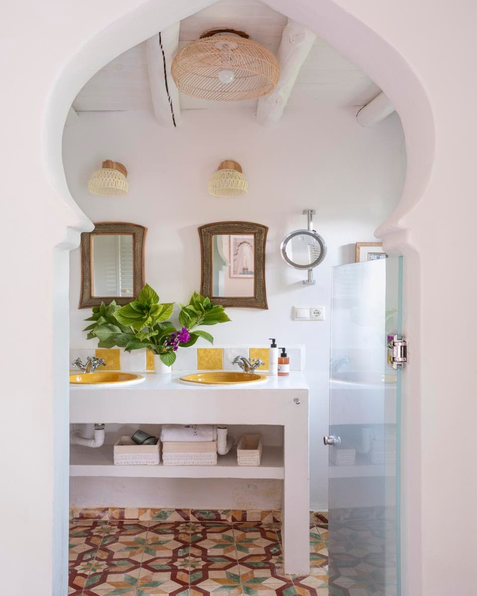 Baño con mueble de lavabo doble en blanco con dos lavabos artesanales amarillos y espejos de marco de mimbre, enmarcado por un arco de yeso blanco. La ducha con azulejos azul marino y el suelo de mosaico hidráulico se aprecian al fondo.