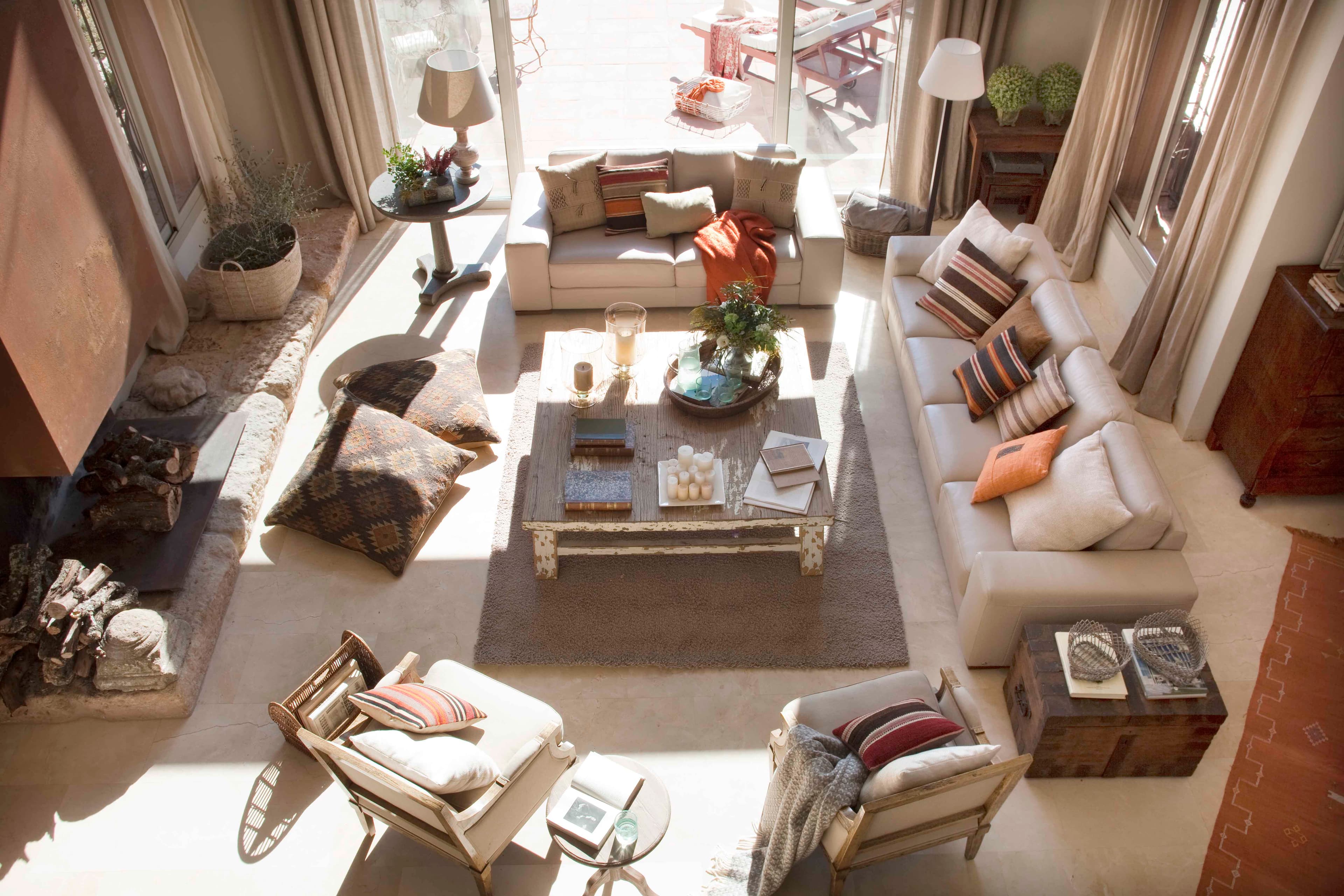 Vista cenital del salón que muestra la distribución completa del espacio en torno a la chimenea de acero corten. Sofás en crudo con cojines de kilim, mesa central de madera envejecida cargada de velas y flores, baúl de madera, sillones de mimbre y una alfombra gris. La luz entra abundantemente por los ventanales hacia la terraza.