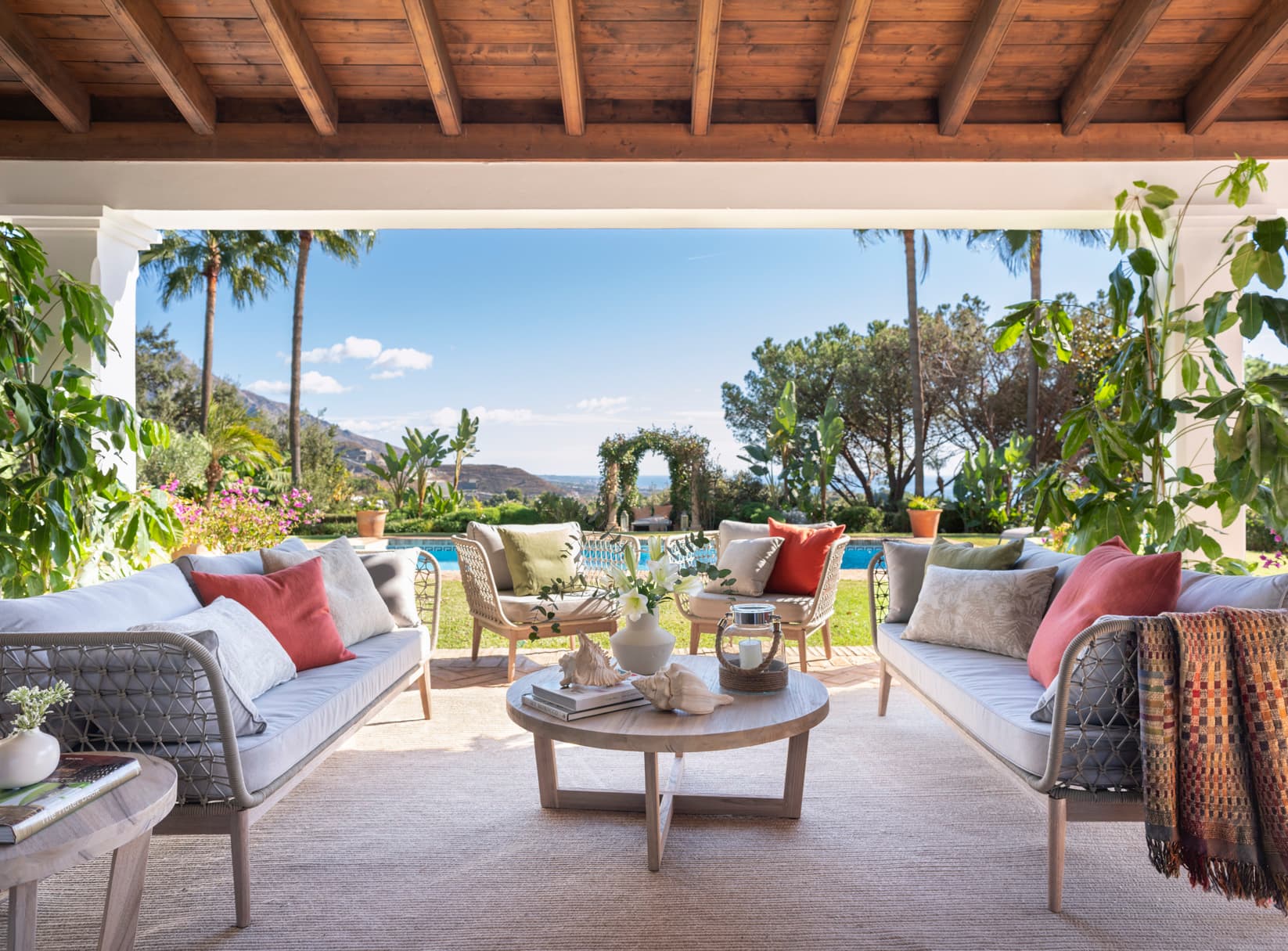 Amplio porche cubierto con techo de madera y vistas espectaculares al jardín tropical, piscina y montañas en el horizonte. Mobiliario outdoor en gris con cojines en tonos tierra, rojo y verde, alrededor de una mesa de centro redonda. Plantas tropicales exuberantes enmarcan la escena con bugambillas al fondo.