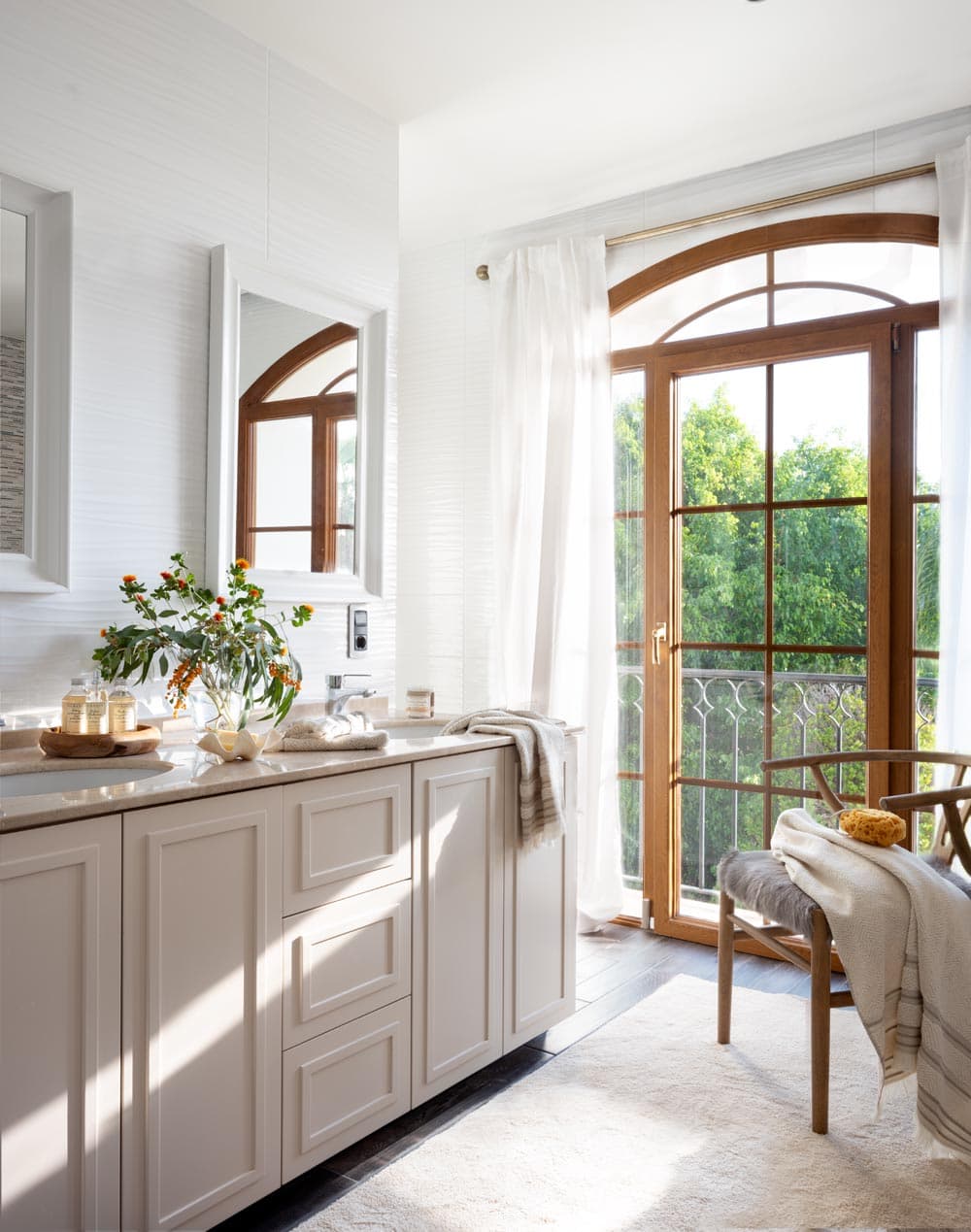 Cuarto de baño luminoso con mueble bajo lacado en blanco roto, encimera de piedra natural, espejo enmarcado en arco y puerta balconera de madera con arco que da a un jardín frondoso. Un jarrón con ramas de piracanta naranja y productos de baño dispuestos añaden un toque natural y sereno.