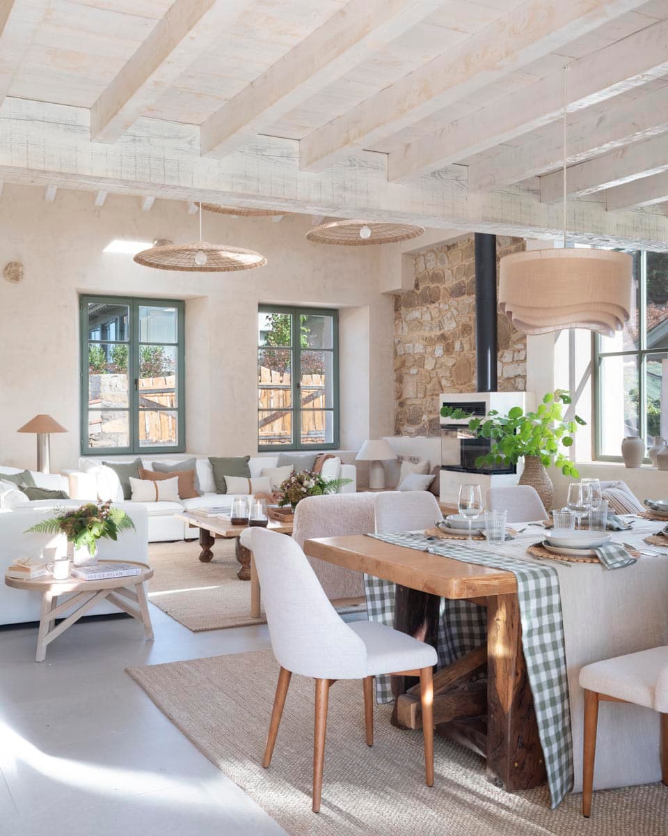 Espacio de planta abierta que integra salón y comedor bajo vigas blanqueadas, con mesa de comedor de madera maciza con camino de cuadros verdes, sillas tapizadas en blanco y lámparas de ratán colgantes.