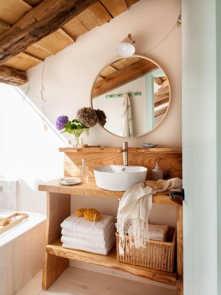 Baño en buhardilla con mueble lavabo de madera natural, lavabo sobre encimera blanco y espejo redondo, vigas rústicas al descubierto y ventana inclinada. Las hortensias moradas, las toallas blancas y la cesta de esparto añaden calidez orgánica al espacio.