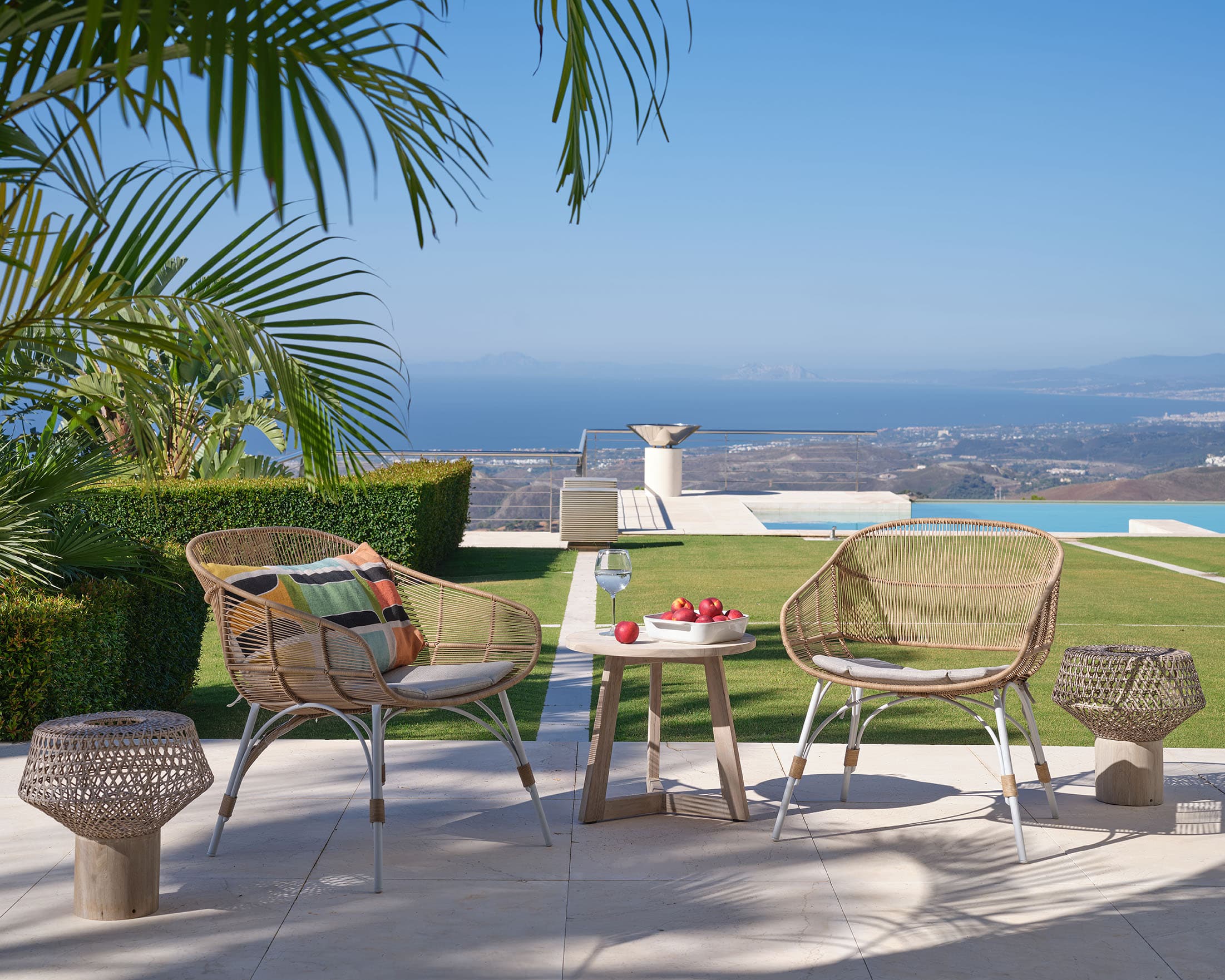 Terraza con sillones de ratán natural y cojines de rayas, pequeña mesa de café de madera, junto a una piscina de borde infinito con vistas a la costa y la ciudad.