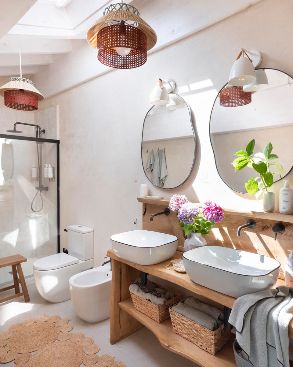 Baño luminoso con mueble de lavabo doble de madera maciza rústica, lavabos cuadrados de sobre encimera en blanco, espejos orgánicos de marco negro, lámparas colgantes de ratán en rojo oscuro y plafón cenital.