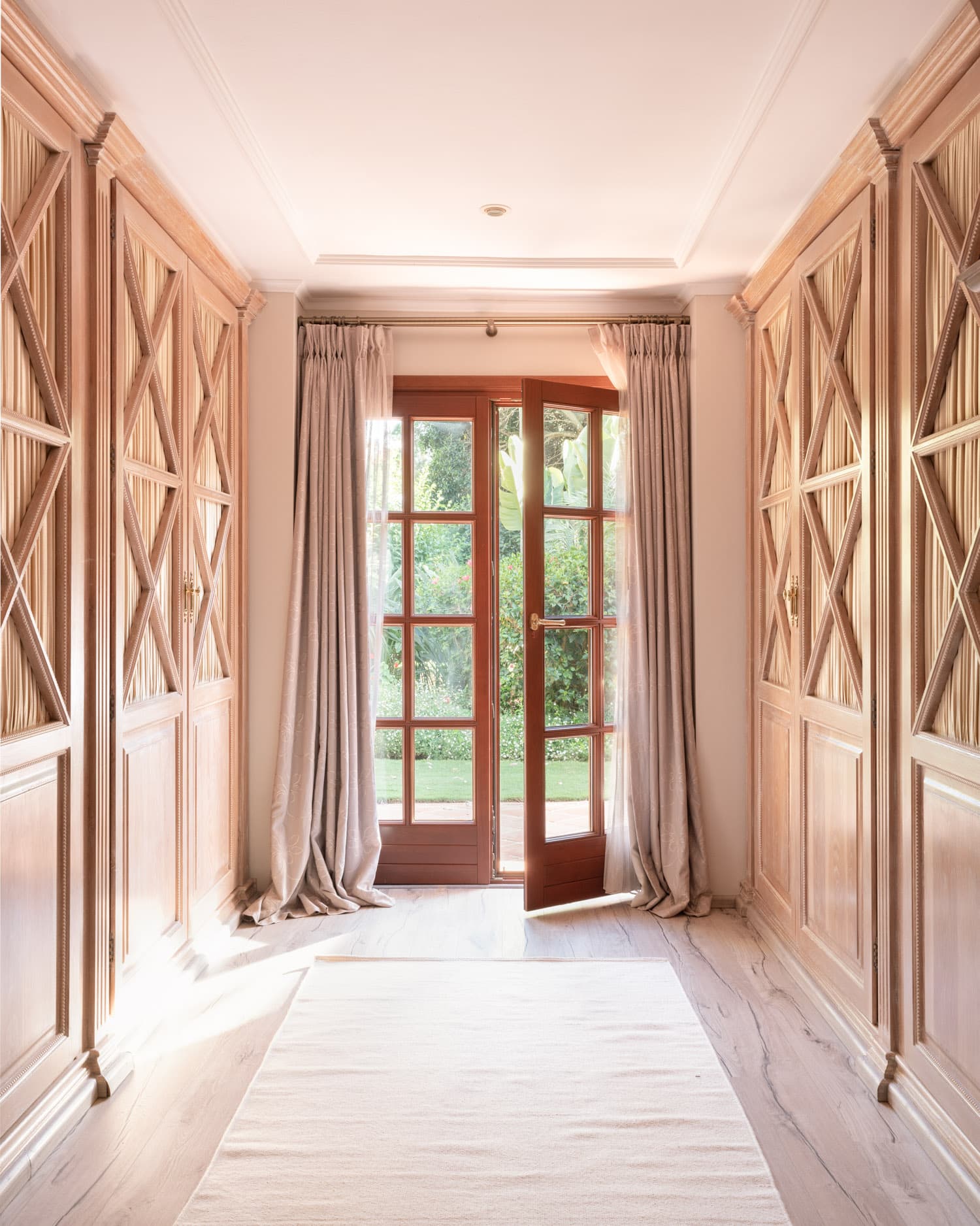 Pasillo elegante revestido a ambos lados con armarios empotrados de madera clara con motivos en X y herrajes dorados, que enmarcan una puerta francesa de madera maciza abierta hacia el jardín. Cortinas de lino en tono arena caen hasta el suelo, y una alfombra blanca recorre el suelo de mármol, inundando el espacio de luz natural.