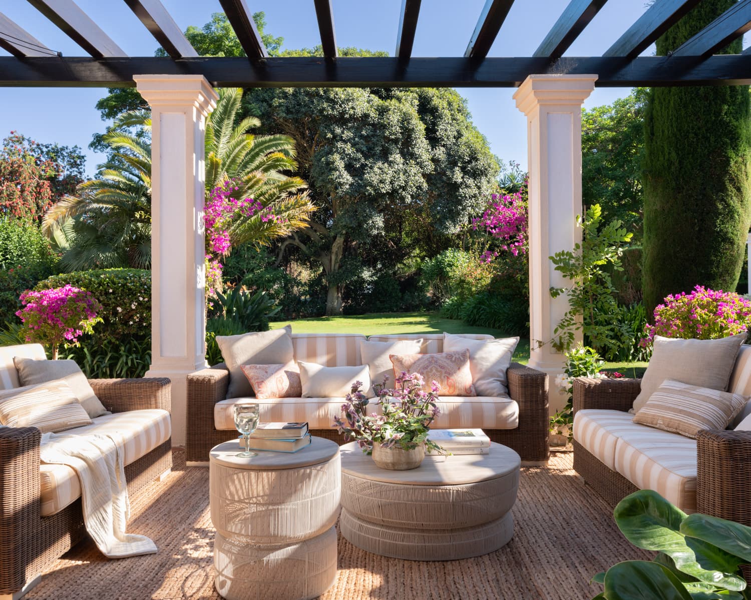 Amplio salón exterior bajo una pérgola con columnas cuadradas blancas. El mobiliario de ratán trenzado con cojines a rayas crema y beige y mesas auxiliares cilíndricas de fibra natural se dispone frente al jardín en plena floración, con buganvillas fucsia, palmeras y cipreses. Una alfombra de yute aporta calidez al suelo de piedra.