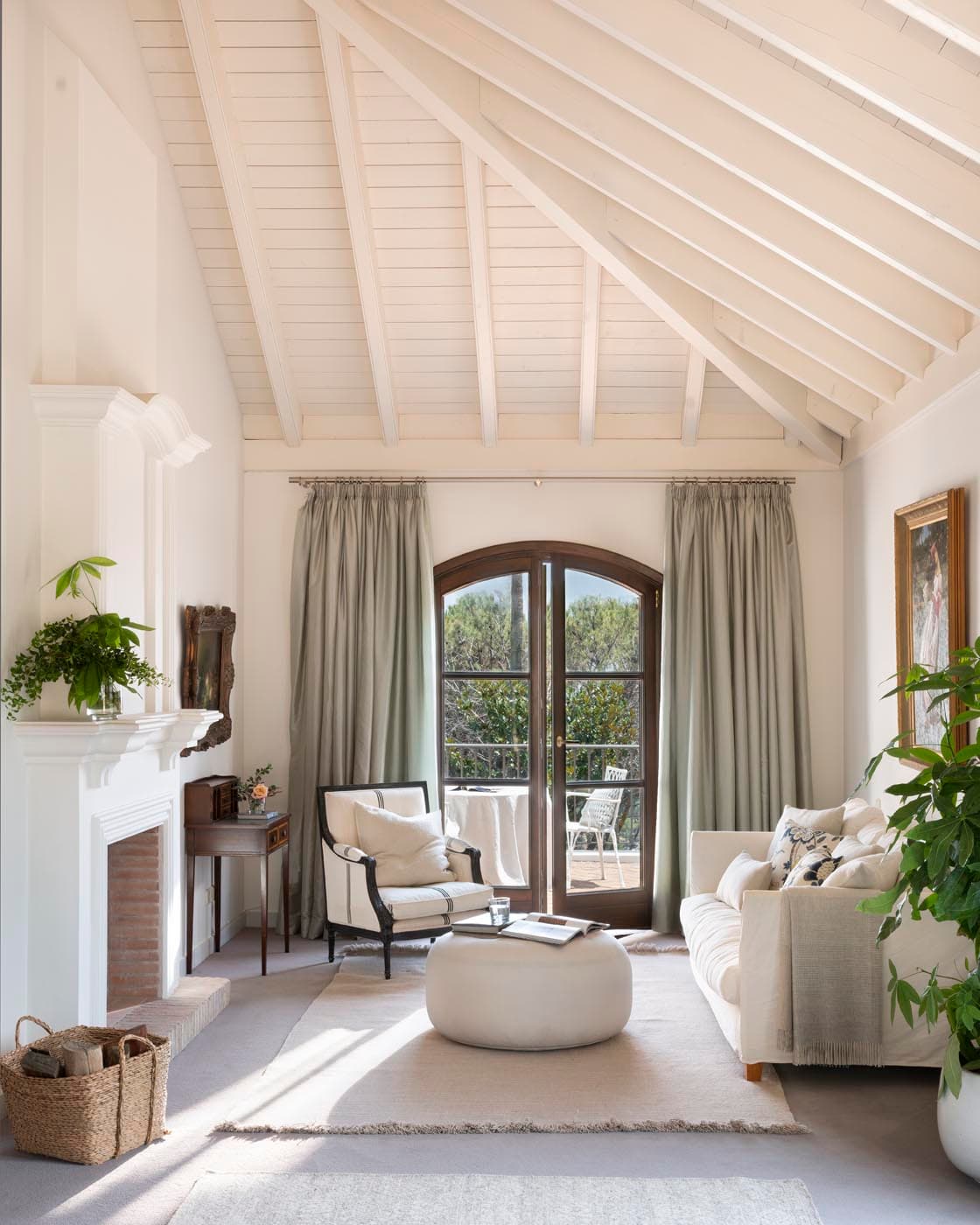 Acogedora sala de estar con techo a dos aguas de vigas y tablillas pintadas en blanco, chimenea blanca, sofá de lino, sillón de estilo Luis XVI en blanco y negro y cortinas verde salvia. Una gran planta tropical y cuadros clásicos de marco dorado complementan el ambiente refinado y sereno.