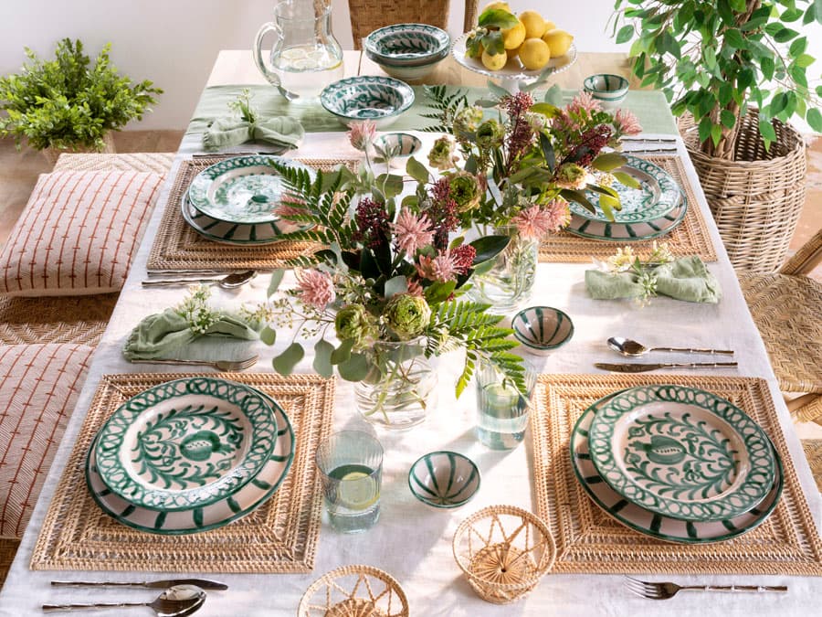 Mesa de comedor exterior vestida con mantel de lino crudo y bajoplatos cuadrados de fibra trenzada. La vajilla cerámica artesanal en blanco y verde botella, con decoración de motivos botánicos, aporta un carácter mediterráneo y artesanal al conjunto. El centro de mesa combina flores silvestres en tonos rosas y burdeos, ramas de eucalipto y helechos, en una jarrita de vidrio transparente. La paleta de colores naturales —crudo, verde, terracota— refleja la esencia del proyecto.