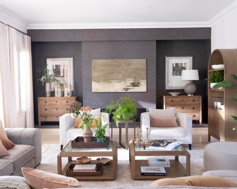 Salón interior de estilo contemporáneo elegante con pared de papel tapiz en gris antracita que contrasta con las paredes blancas. Dos sillones blancos simétricos, cómodas de madera natural, estantería de madera arqueada con objetos decorativos y sofás de color gris. Cuadros abstractos y plantas añaden sofisticación al conjunto.