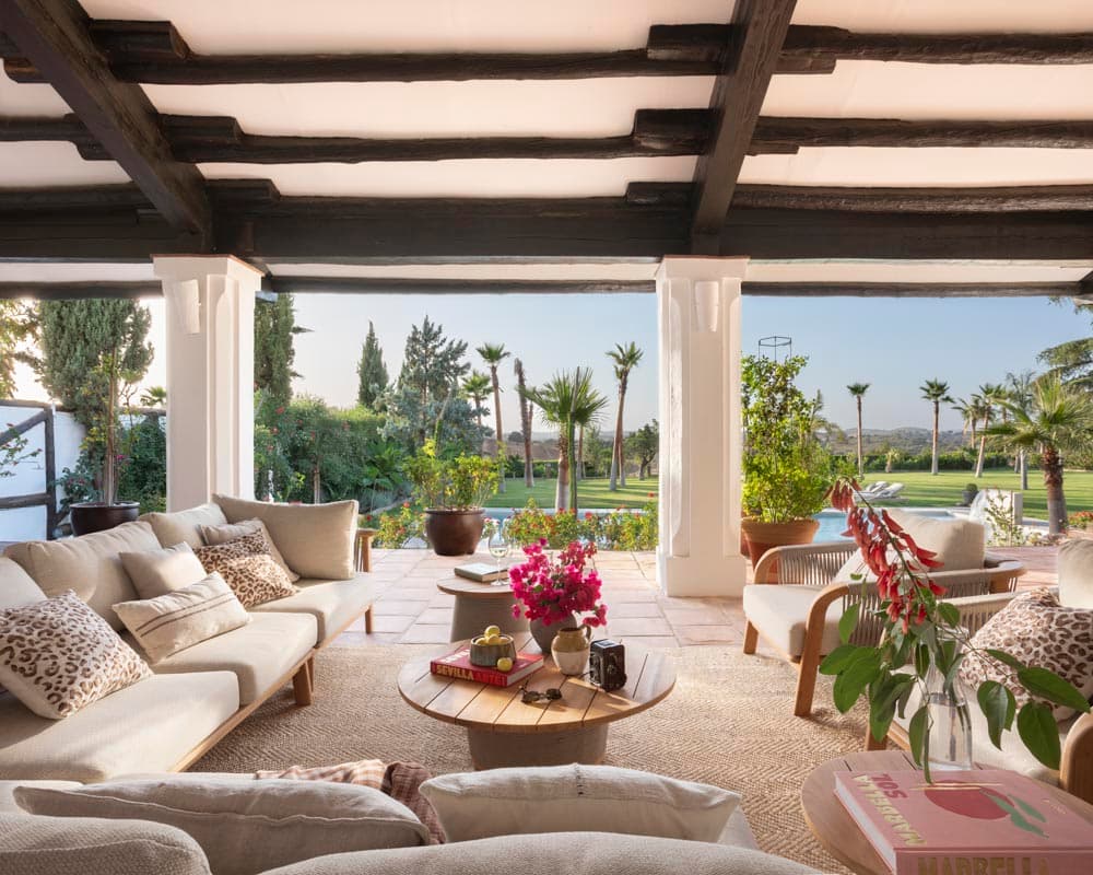 Zona de estar del porche exterior con sofás blancos de lino y sillones de madera con cojines de estampado leopardo, mesa redonda de madera con buganvilla. Vigas de madera negra y vistas al jardín con palmeras y piscina.