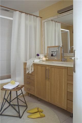 Cuarto de baño sencillo y luminoso con mueble de lavabo en madera de roble natural, espejo retroiluminado, cortina de ducha de lino blanco y taburete industrial de metal. Los detalles en tonos ocre, como las zapatillas amarillas y el marco de fotografía, añaden calidez al ambiente nórdico funcional.