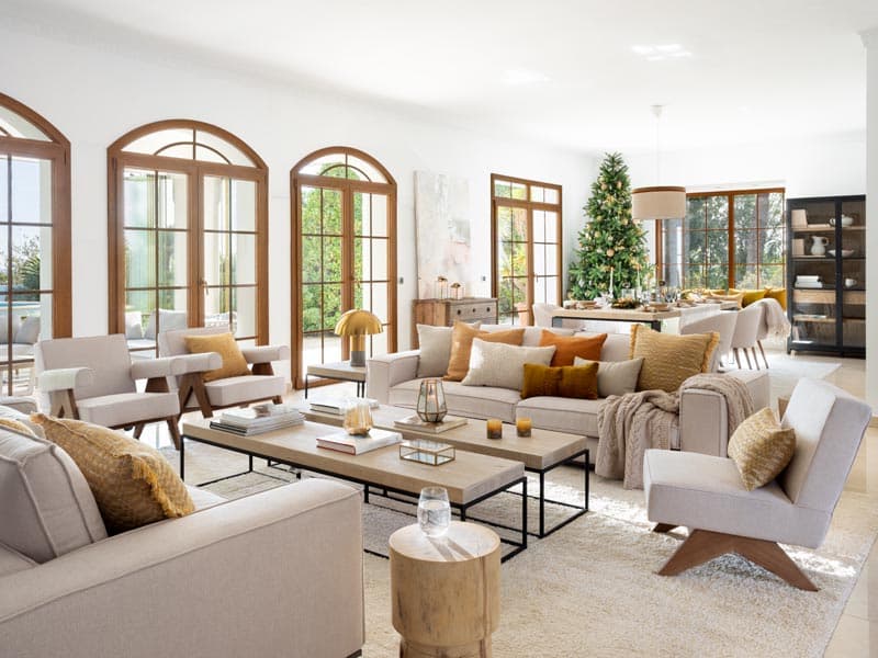 Vista amplia del salón-comedor con árbol de Navidad decorado con adornos dorados al fondo, sofás y sillones crema con cojines en cobre y mostaza, mesa de coffee con troncos y velas, y la vitrina negra en el límite de la zona de comedor. La decoración festiva enriquece el espacio sin romper la elegancia del proyecto.