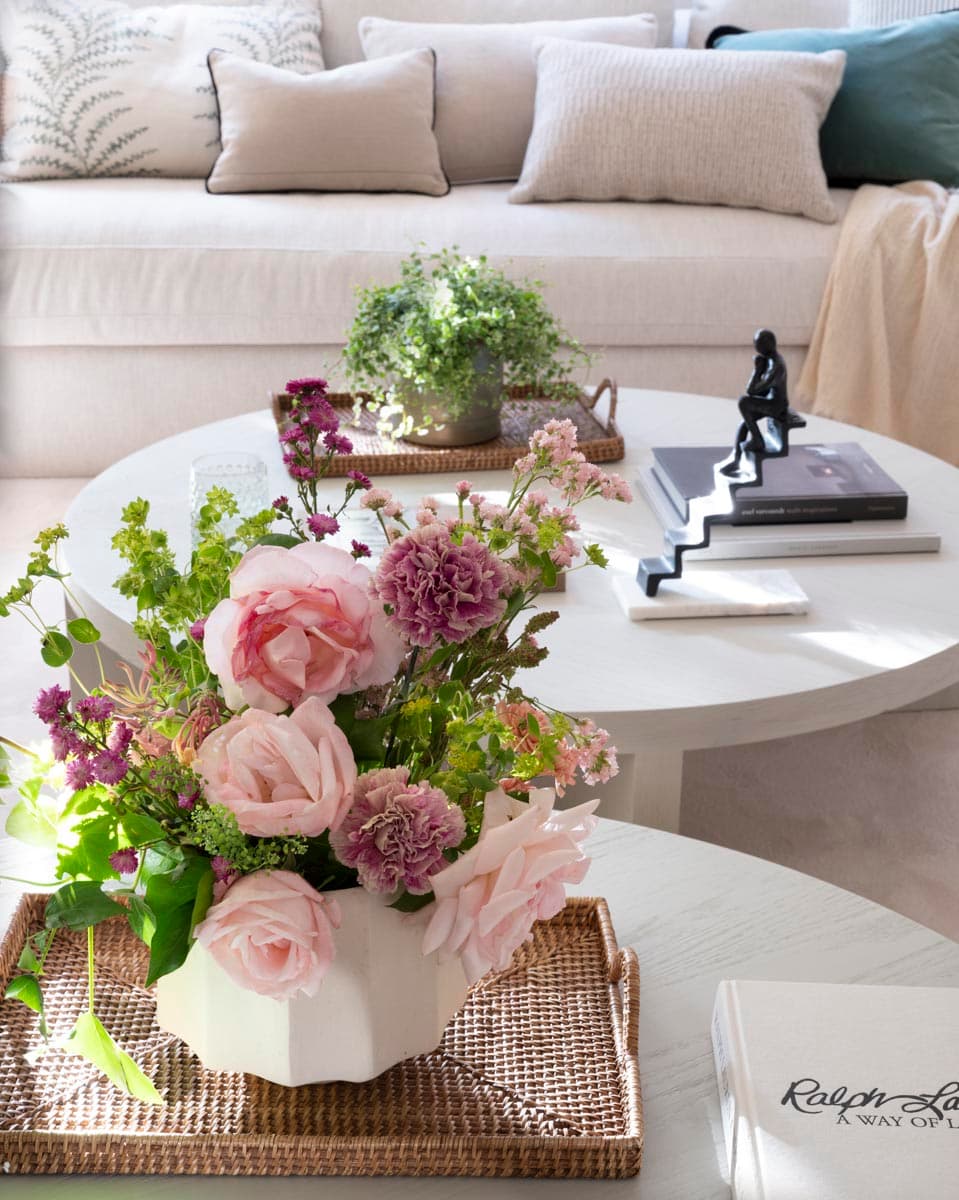 Primer plano de la mesa de centro redonda blanca con un ramo de rosas rosas y claveles malva en jarrón cerámico blanco sobre bandeja de mimbre, acompañado de libros de decoración y una escultura negra. Al fondo, los cojines con bordado de helecho sobre el sofá blanco añaden profundidad a la composición.
