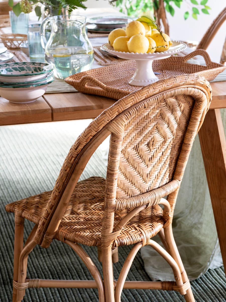 Primer plano de una silla de ratán con respaldo y asiento en tejido de espiga natural muy detallado. Sobre la mesa de madera al fondo se aprecia una bandeja de ratán con una fuente blanca de pedestal con limones, vasos de agua y otros elementos de vajilla artesanal. La calidad del trabajo artesanal del ratán y la calidez de la madera son protagonistas de este plano de detalle.