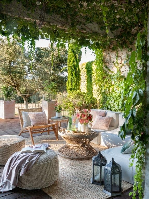 Vista más abierta del porche exterior cubierto de hiedra y vegetación con sillón bajo de madera y ratán, sofá blanco con cojines, puf de fibra y faroles marroquíes. Al fondo, una pared de piedra natural y jardín con cipreses y vistas al campo abierto con iluminación cálida de tarde.