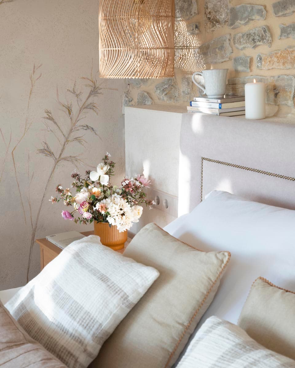 Detalle de la mesita de noche con ramo de flores silvestres en macetero ocre estriado, cabecero tapizado y fondo de mural botánico pintado sobre enlucido beige junto a muro de piedra vista.