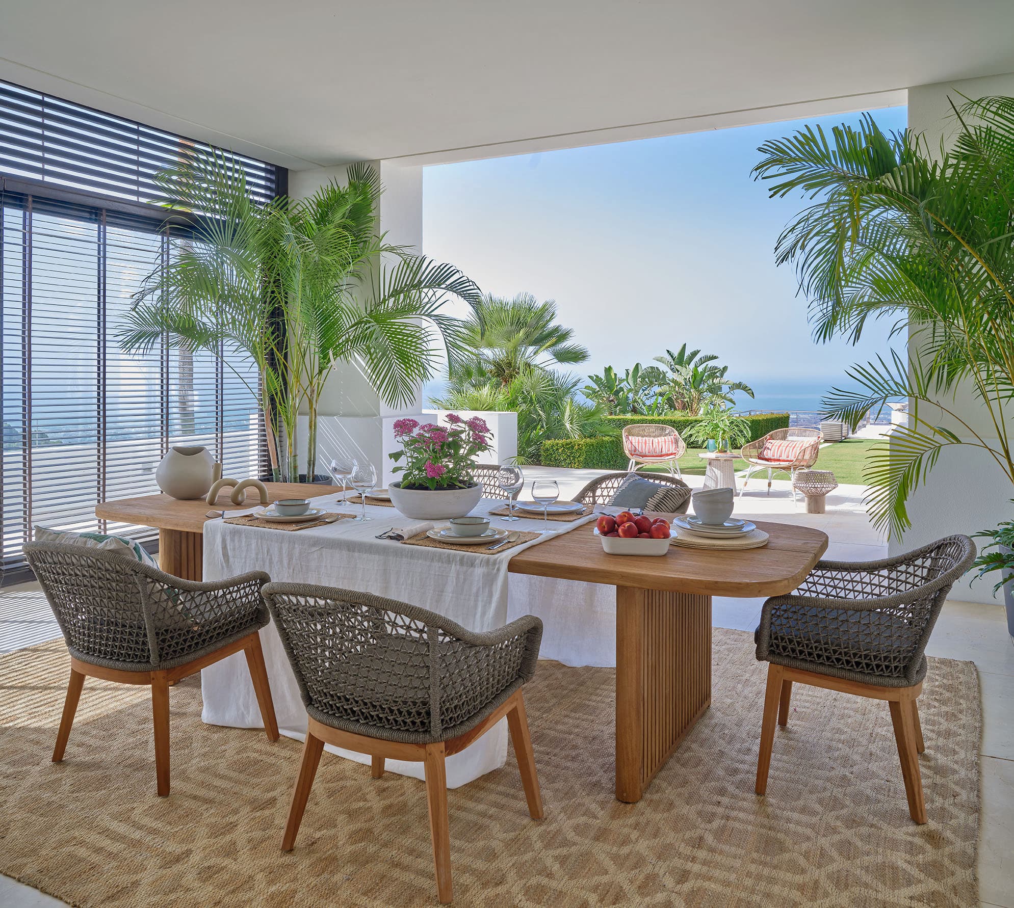Comedor exterior cubierto de una villa en La Zagaleta, con mesa de madera de teca, sillas de cuerda trenzada y vistas panorámicas al Mediterráneo y las palmeras del jardín.