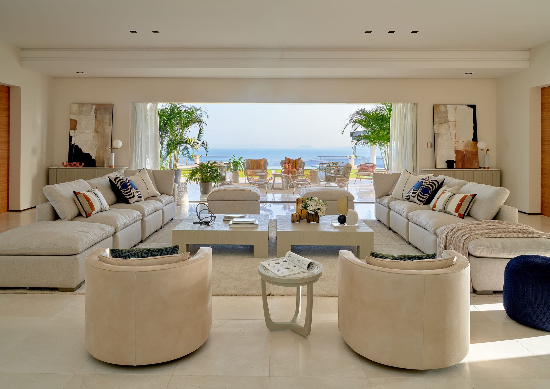 Salón principal de gran amplitud con sofás modulares en beige claro, sillones curvos, mesas de café lacadas blancas y una espectacular apertura hacia la terraza con vistas al mar.