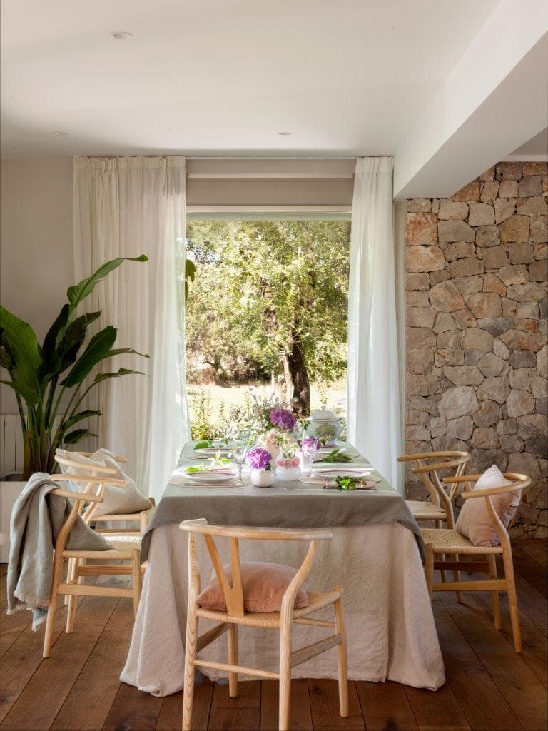 Comedor con pared de piedra natural vista a la derecha y gran ventanal que enmarca el jardín. Mesa vestida con mantel de lino bicolor y sillas tipo Wishbone de madera clara. La gran planta estrelicia aporta un toque tropical. Las flores rosas y moradas en jarrones blancos y una manta de punto en el respaldo completan el ambiente.