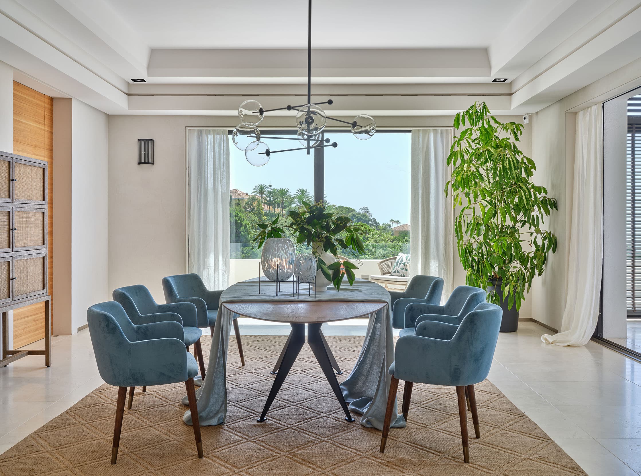 Comedor con mesa redonda de madera sobre base metálica negra, sillas en terciopelo azul, una gran planta interior y lámpara de esferas de cristal, con acceso directo al jardín.