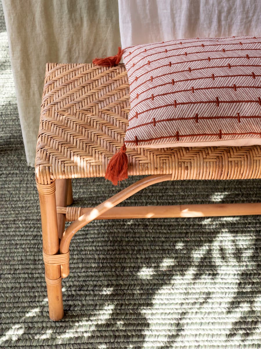 Detalle de un banco de ratán tejido con patrón en espiga sobre una alfombra de yute con dibujo geométrico en verde y crudo. Sobre el banco, un cojín rectangular de lino crudo con estampado de líneas en terracota y borlas en los extremos. La luz natural crea un juego de sombras sobre la alfombra y el banco, enfatizando la textura de los materiales naturales.