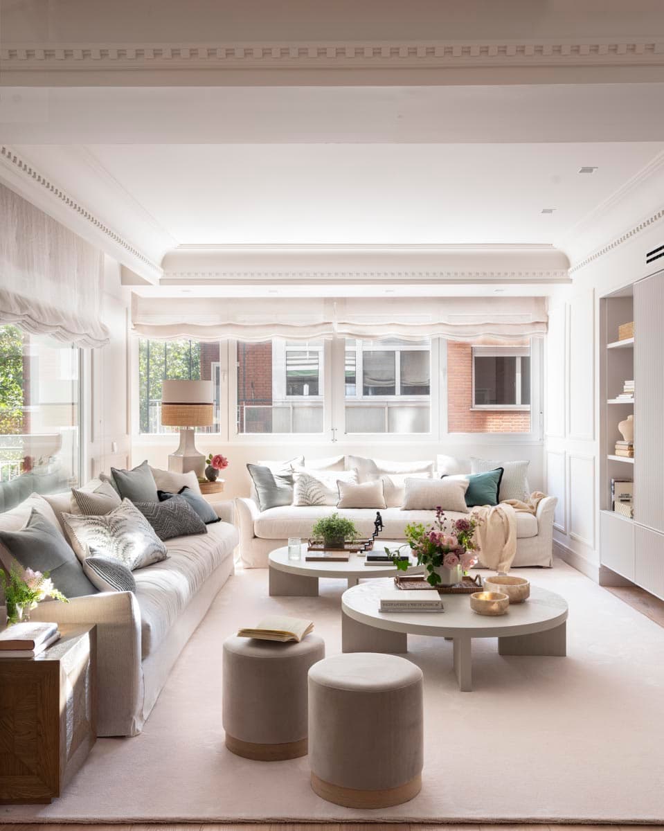 Gran salón luminoso con dos sofás largos de lino blanco con cojines en azul pizarra, gris y verde, dos mesas de centro redondas en gris cemento y puf redondos de terciopelo gris en primer plano. La gran librería blanca a la derecha, las molduras en techo y los estores con volante de lino enmarcan un espacio de estética clásica contemporánea.