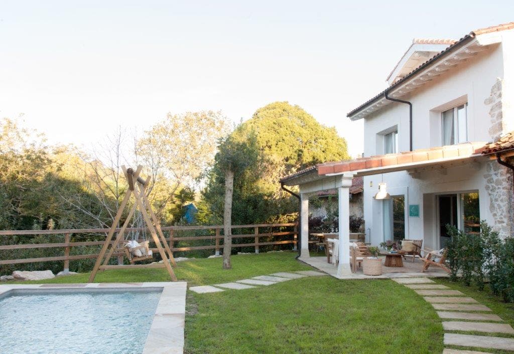 Vista exterior de la casa con fachada blanca y tejado de teja, jardín cuidado con piscina en primer plano y columpio de madera. El porche cubierto con mobiliario exterior y la vegetación rodean la vivienda de estilo rural contemporáneo.