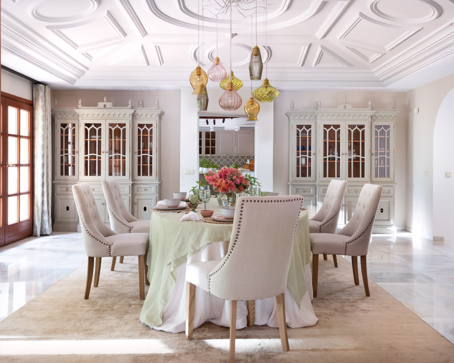 Comedor formal de gran altura con un espectacular techo de molduras geométricas en blanco. Un conjunto de lámparas colgantes de cristal soplado en tonos ámbar, rosa, verde y gris ceniza forman una instalación artística sobre la mesa ovalada. Cuatro vasos de vitrina estilo neoclásico gris verdoso a juego flanquean la sala, mientras sillas tapizadas en lino con tachuelas enmarcan la mesa.