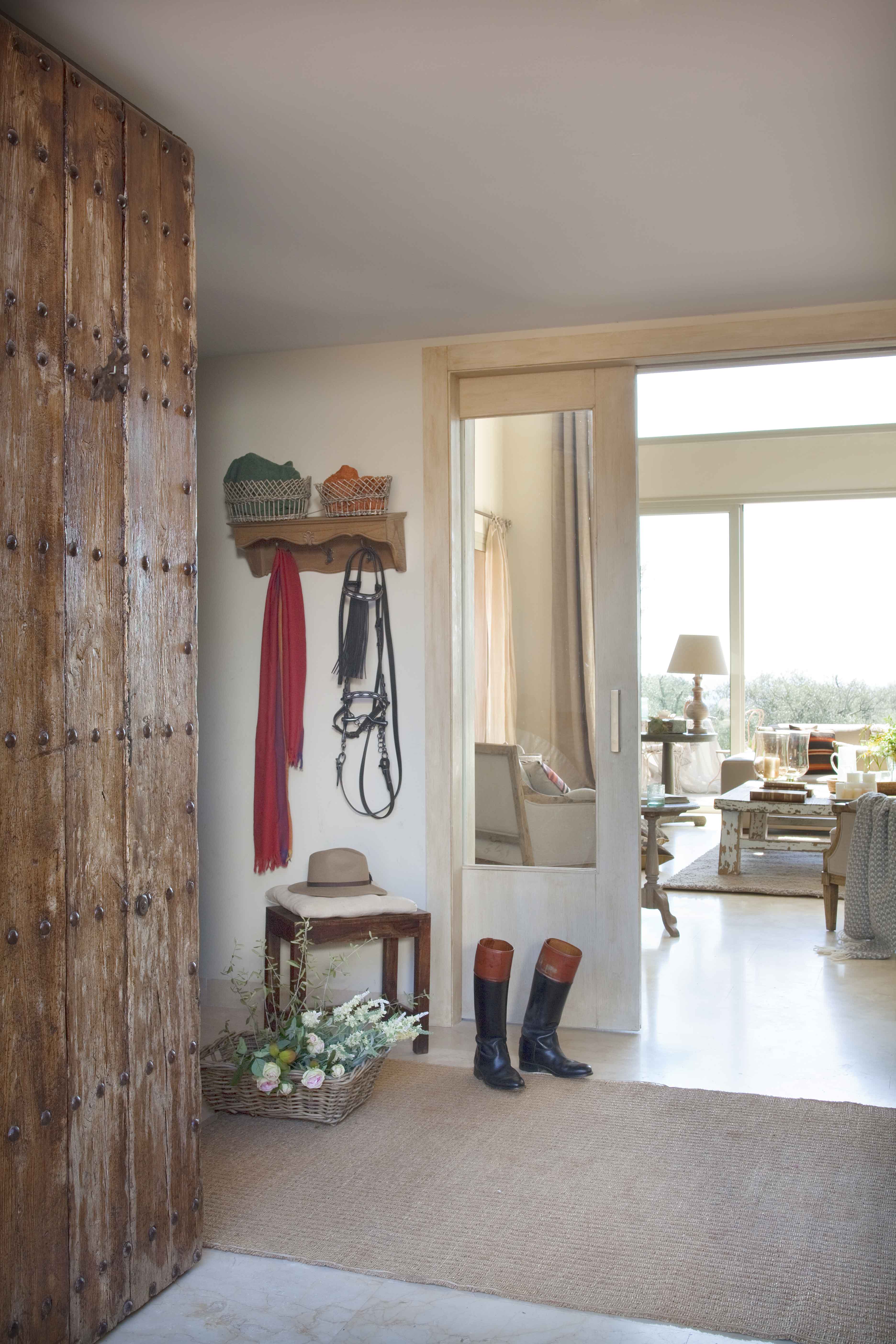 Recibidor donde una gran puerta de madera tachonada con apliques de hierro forjado marca la entrada. En la pared, un perchero rústico con riendas de caballo, bufandas y sombreros. Botas de montar apoyadas en el suelo junto a una cesta de flores. Al fondo, el luminoso salón con vistas al campo.