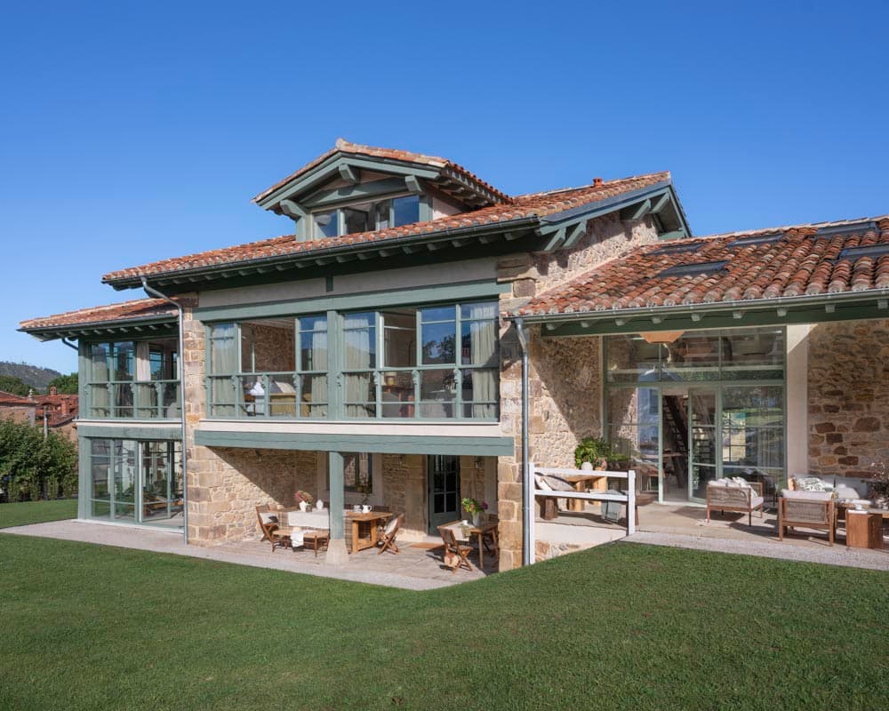 Vista diurna de la fachada principal con galería acristalada de dos alturas en verde, terraza pavimentada con mobiliario de madera, estructura de piedra vista y cubierta mixta de teja árabe y lucernarios.