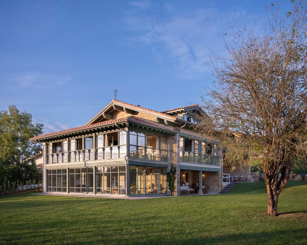 Fachada trasera de la casa rehabilitada al atardecer, con estructura de piedra original, galerías acristaladas de marco gris-verde, tejado de teja tradicional y gran jardín en primer plano.