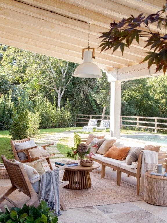 Terraza cubierta con sofá y sillones de madera natural con cojines en crudo y mostaza, mesa de café con estructura acanalada y vistas a la piscina y jardín. La lámpara industrial blanca colgante y el árbol de hoja roja aportan contraste al espacio exterior.