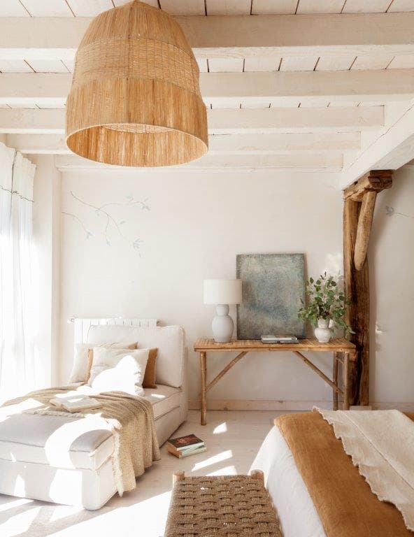Dormitorio aireado con viga de madera rústica como elemento estructural, sofá cama individual en lino blanco y lámpara colgante de rafia grande. Al fondo, una mesa consola de madera con cuadro abstracto y detalles decorativos naturales.