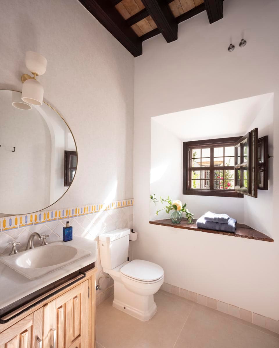 Baño con cenefa artesanal de azulejo en azul y amarillo, espejo redondo de latón y aplique doble de latón. Lavabo de piedra empotrado en mueble de madera, ventana de madera con alféizar de madera y flor en jarrón. Vigas oscuras en el techo.