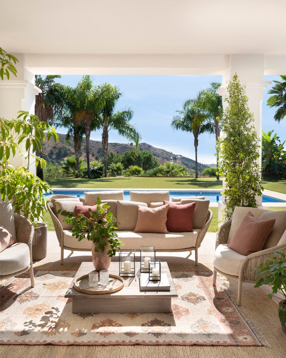 Zona de estar exterior bajo porche con sofá curvo de ratán y tejido en beige con cojines terracota y rosa. Mesa de centro de piedra con farol y objetos decorativos. Las palmeras, la piscina azul y las montañas al fondo crean un entorno de lujo típico de la Costa del Sol.