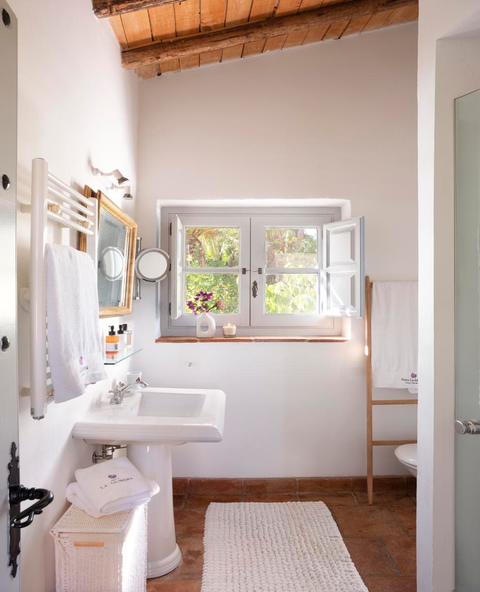 Baño sencillo con lavabo de pedestal blanco, espejo dorado enmarcado y toallas bordadas con el logo de La Morera. La ventana con contraventanas grises da al jardín y una escalera de madera porta toallas adicionales.
