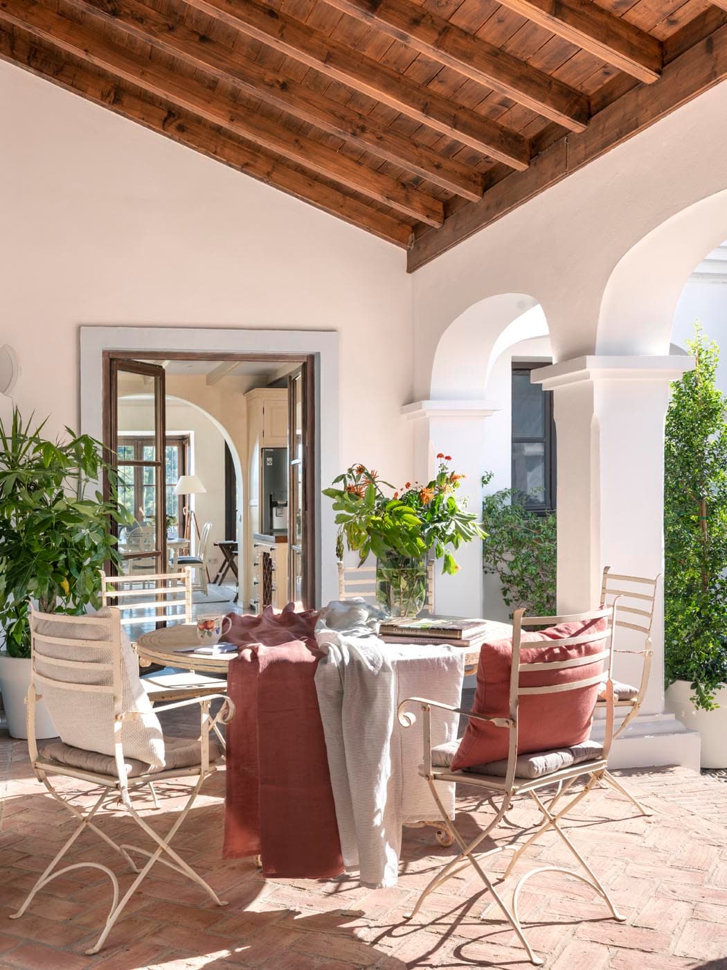 Terraza cubierta con techo de vigas de madera, suelo de barro en espiga y sillas plegables de hierro forjado alrededor de una mesa con centro floral, delimitada por arcos blancos con vistas al jardín. Los cojines en terracota y gris aportan un toque de color cálido y desenfadado.