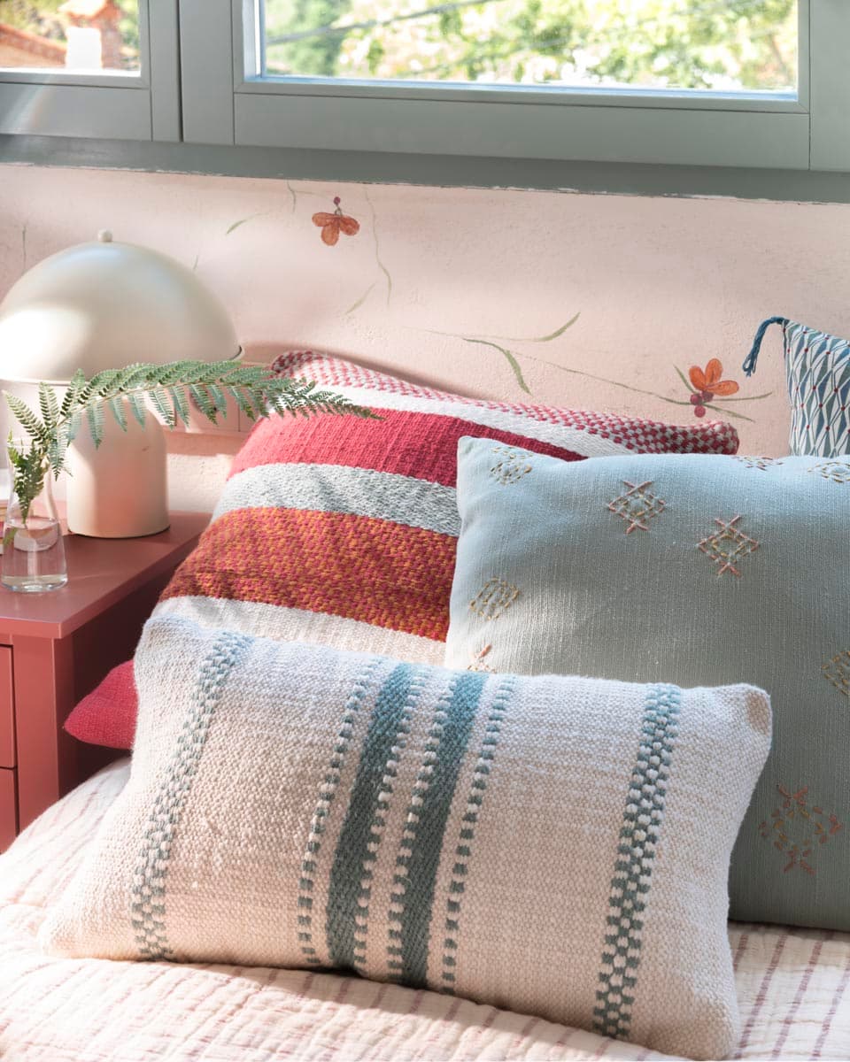 Detalle de cama infantil con cojines en colores vivos: rayas de rojo y naranja, punto en azul celeste con motivos romboidales y blanco con listas azules, sobre fondo de mural pintado con pequeñas flores.