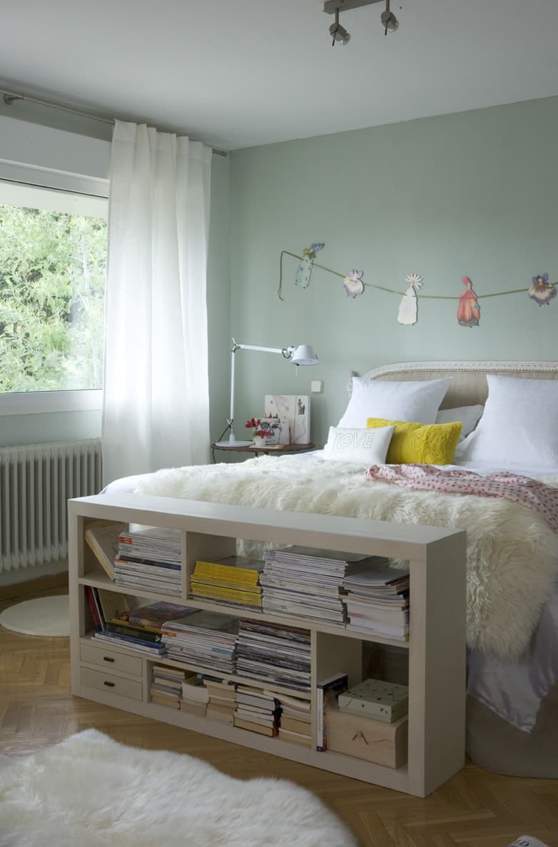 Dormitorio de tonos suaves con paredes en verde menta, cama tapizada en lino con ropa de cama blanca y cojín amarillo mostaza. Una estantería baja blanca a los pies de la cama almacena revistas y libros. Una guirnalda de ilustraciones florales decora la pared del cabecero. Suelo de parqué y alfombra blanca de pelo largo.