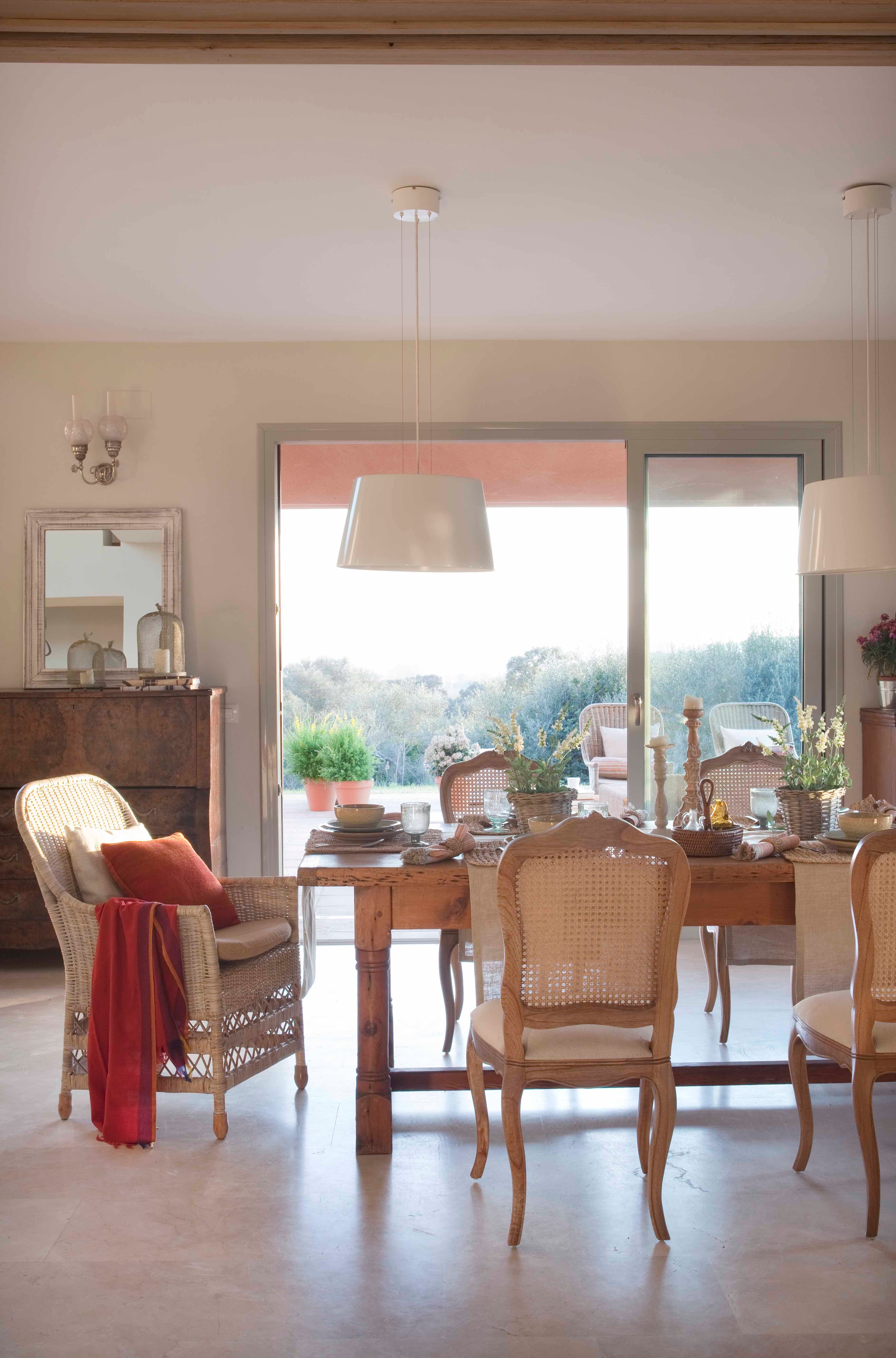 Comedor luminoso con mesa de madera rústica y sillas de rejilla de estilo francés. Una lámpara colgante blanca de gran formato ilumina el espacio que se abre al jardín a través de amplias puertas correderas. En el fondo, un aparador antiguo con espejo y vajilla. Los detalles florales y la manta terracota añaden calidez.
