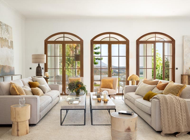 Salón equilibrado con tres arcos de madera que enmarcan el jardín exterior, sofás simétricamente dispuestos en beige arena con cojines dorados y una manta de punto en el reposabrazos.